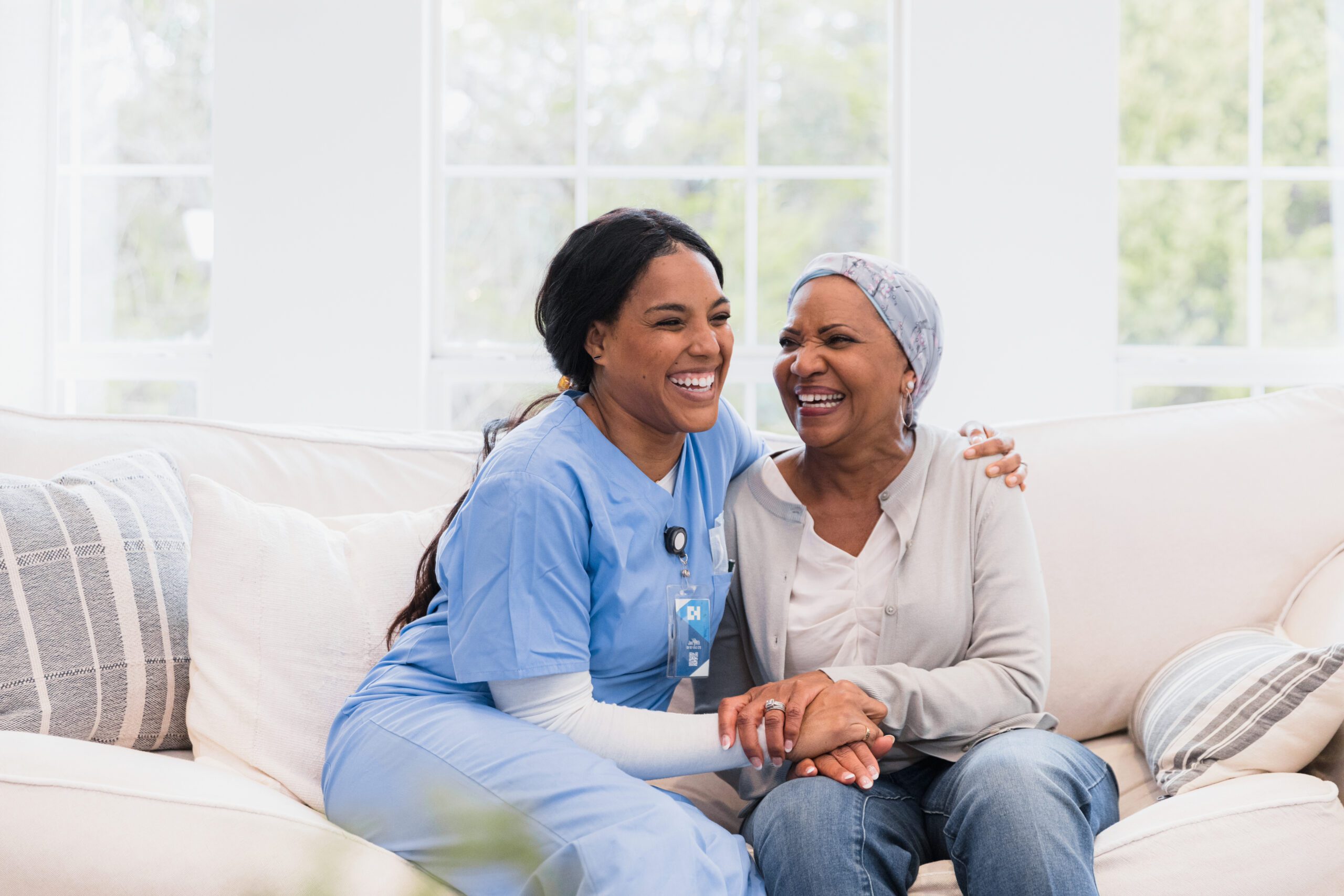 During her visit, the mid adult home health nurse and the senior adult woman with cancer embrace and laugh together. |
