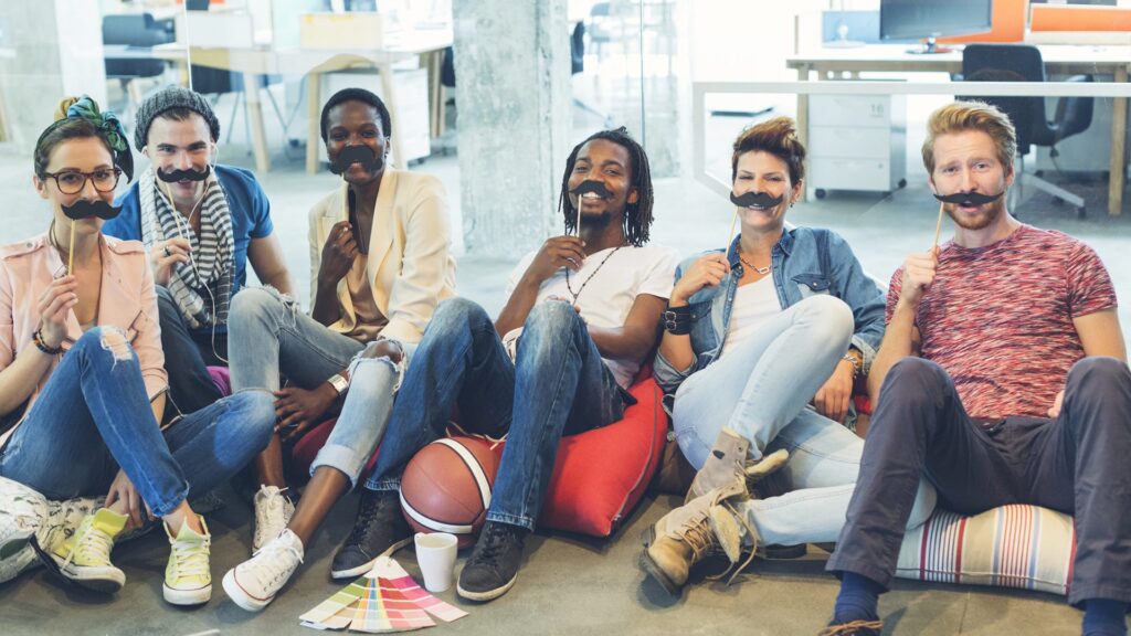 Happy smiling multi ethnic group of designer sitting on floor and holding fake mustache as prostate cancer awareness day #Movember. Looking at camera and smiling.