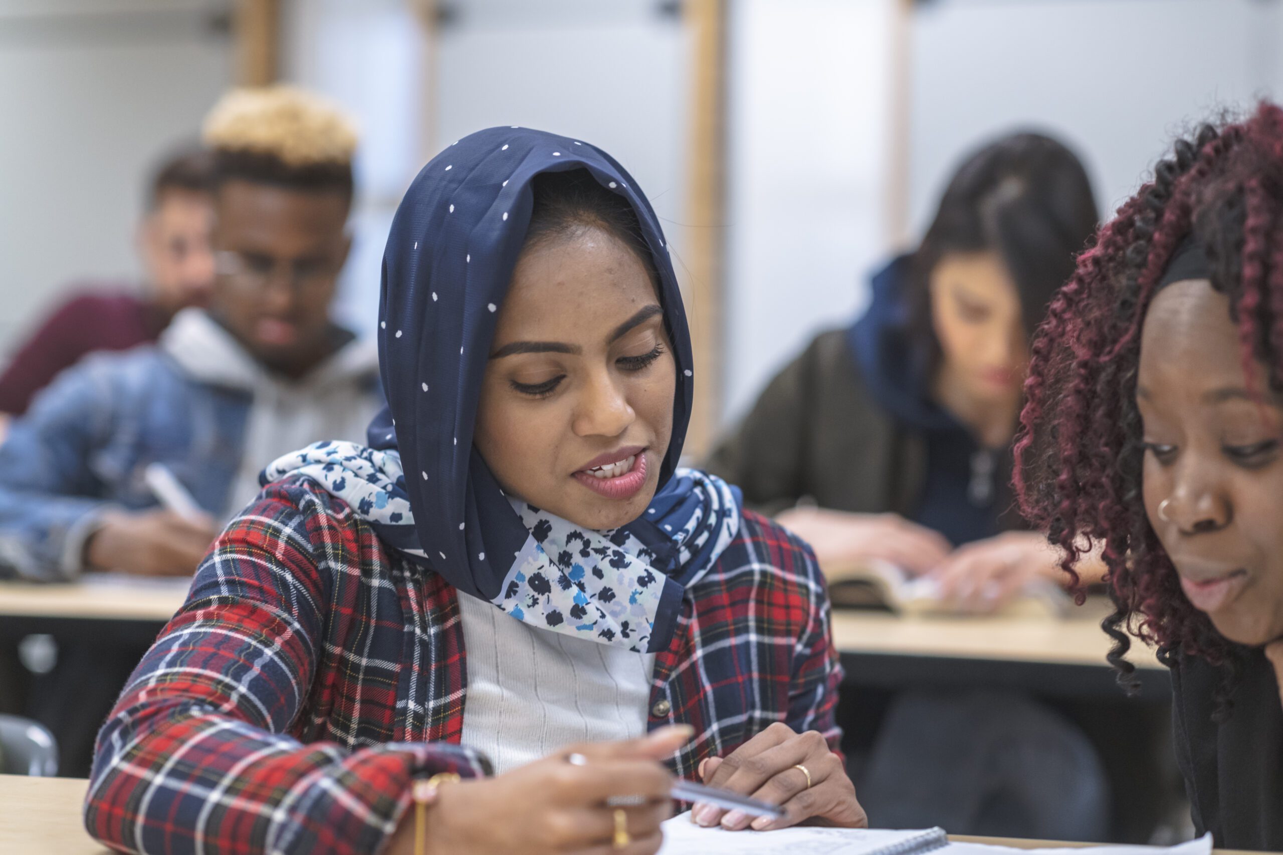 A multi-ethnic group of students sits in a lecture hall listening to their professor off-screen. They are taking notes with the focus being on a female of Indian descent wearing a hijab.