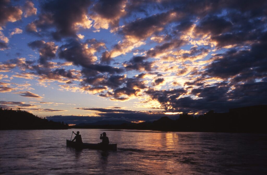 Two men paddle a canoe down the Yukon River near Whitehorse, Yukon, Canada at midnight close to summer solstice. | Deux hommes pagayent en canot sur le fleuve Yukon près de Whitehorse, au Yukon, au Canada, à minuit, à l'approche du solstice d'été.