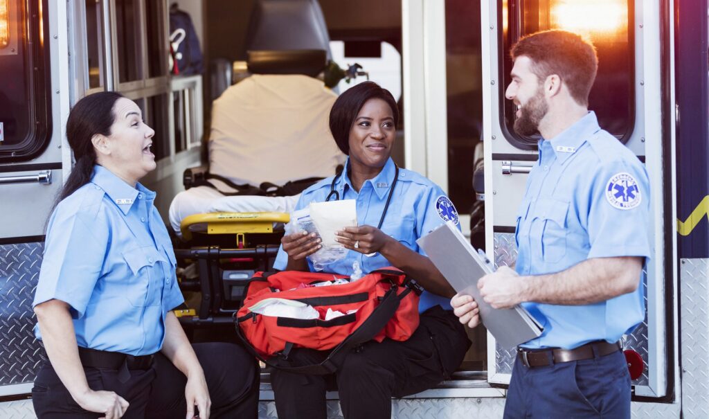 A multi-ethnic group of three paramedics hanging out at the rear of an ambulance, in front of the open doors, conversing.  The focus is on the mature Hispanic woman on the left and the young African-American woman in the middle.