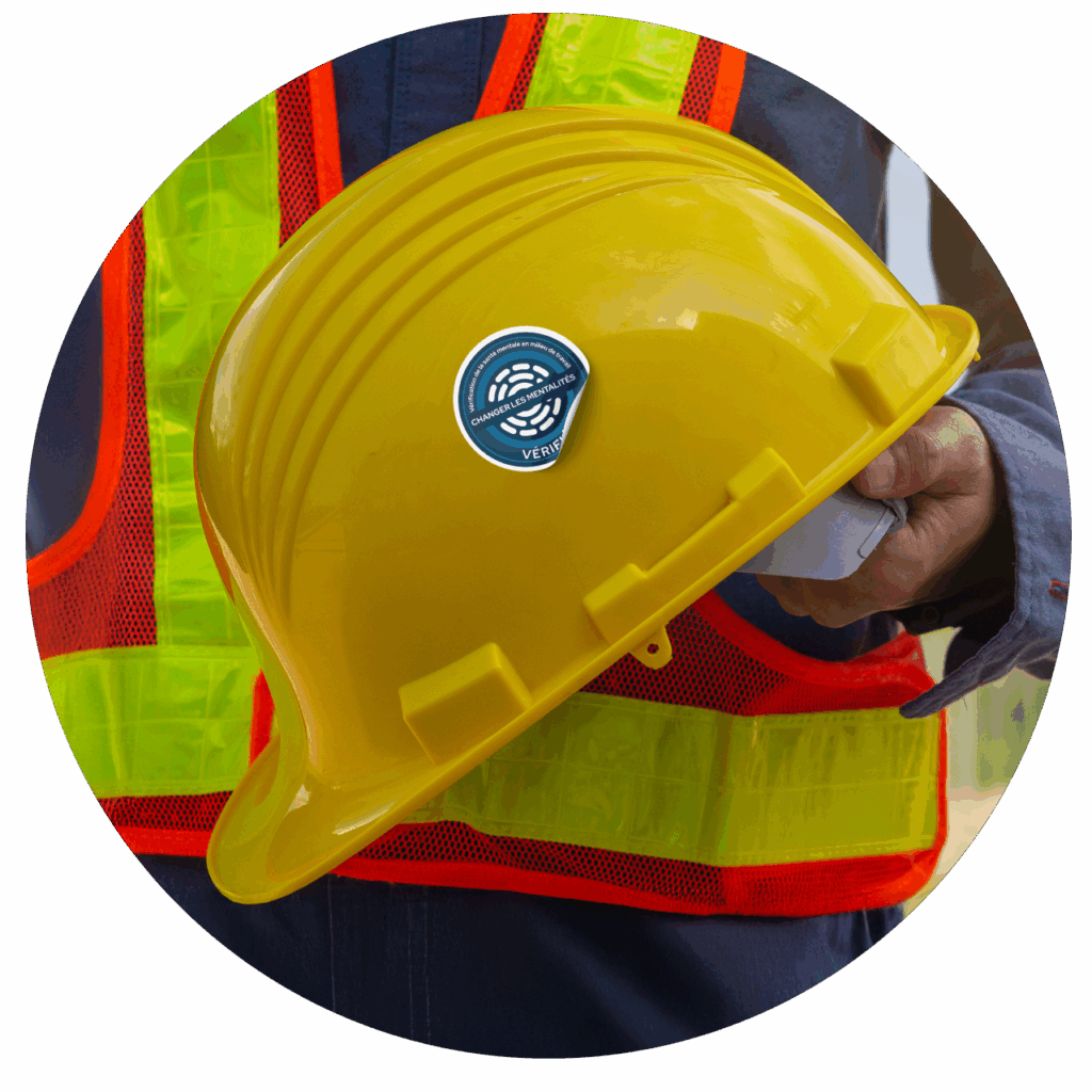 Un homme tient un casque de sécurité jaune avec un badge bleu, indiquant son rôle dans un contexte de construction ou de sécurité.